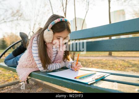 Piccolo splendido artista Disegno con matite colorate, ragazza seduta su una panchina nel soleggiato autunno park, ora d'oro Foto Stock