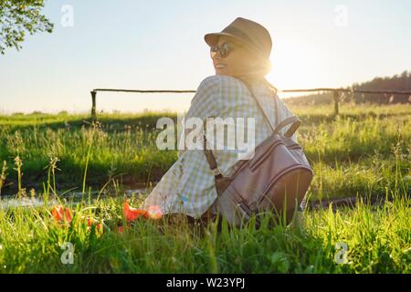 Donna adulta in hat con zaino seduta indietro, estate sfondo del paesaggio, Prato al tramonto, ora d'oro Foto Stock