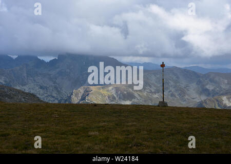 Segnale sul crinale di Zeleni Rila, montagna di Rila, Bulgaria; tempo tempestoso con cielo drammatico; a distanza - cima di Malyovitsa nascosta nelle nuvole Foto Stock