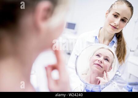 Donna matura guardando in uno specchio mentre medico donna in piedi dietro a. Foto Stock