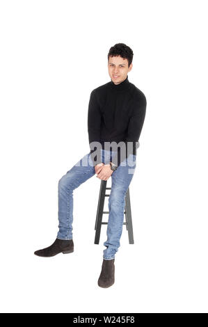 Bel giovane in un maglione nero e jeans seduti su una sedia bar sorridente nella fotocamera, isolato per sfondo bianco Foto Stock