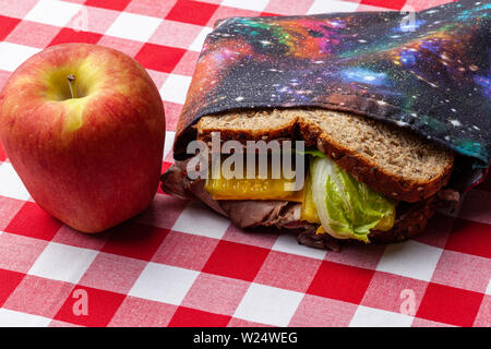 Una mela e un panino in una rispettosa dell'ambiente, in casa, spazio riutilizzabile a tema borsa sandwich seduti su un rosso e bianco tovaglia a scacchi Foto Stock