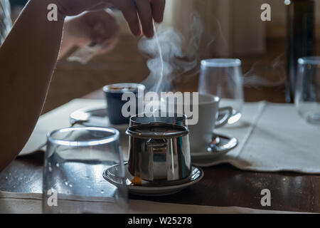 Piccola teiera di metallo con un sacchetto da tè, alcuni bicchieri sul ristorante tavolo. Bianco vapore aumenta al di sopra di acqua calda. Messa a fuoco selettiva. Foto Stock