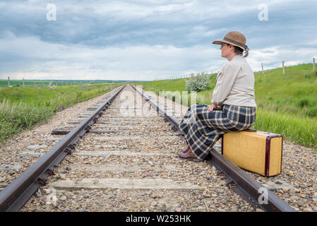 Giovane donna in abiti storici con gli occhiali seduto sulla valigia vintage sulla ferrovia via guardando avanti- viaggio concetto Foto Stock