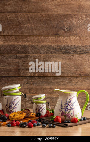 Latte in caraffa in ceramica e due vasi di dimensioni con biscotti e mix di frutti di bosco su sfondo di legno Foto Stock