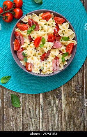 Gustosi piatti di pasta farfalle con salsicce alla griglia, freschi pomodori ciliegini e basilico su una piastra su un sfondo di legno. Vista superiore, piatto laici. Foto Stock