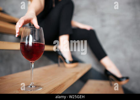 Giovane donna ha problemi con alcool. Vista di taglio del modello raggiungendo il lato di vetro con il vino rosso. Seduta sui passi da solo. Nero elegante abito femminile Foto Stock