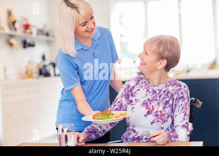 Femmina assistente di cura che serve pasti a Senior donna seduta in carrozzella a tavola Foto Stock