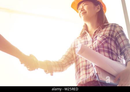 Felice donna asiatica ingegnere come lavoratori edili con blueprint e contratto in mano con protezione Casco giallo si stringono la mano durante il lavoro Foto Stock