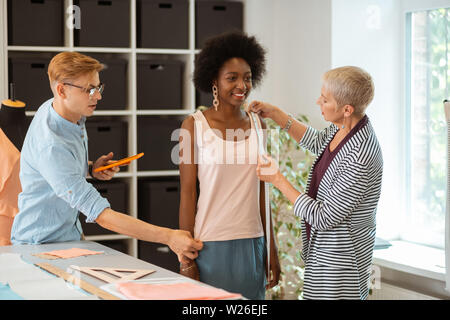 Prendendo misurazioni di abbigliamento. Modello di felice in piedi in compagnia di due stilisti di moda tenendo la sua misurazioni Foto Stock