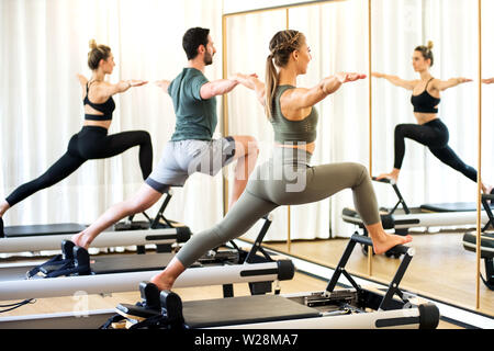Classe in una palestra facendo pilates affondi permanente su letti di reformer di allungare e tonificare la muscolatura riflessa in uno specchio a muro Foto Stock