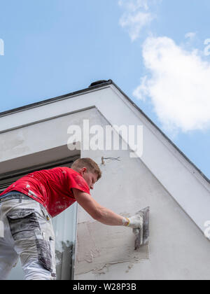 Stuccatore in maglietta rossa funziona su intonaco bianco della vecchia casa durante lavori di isolamento Foto Stock