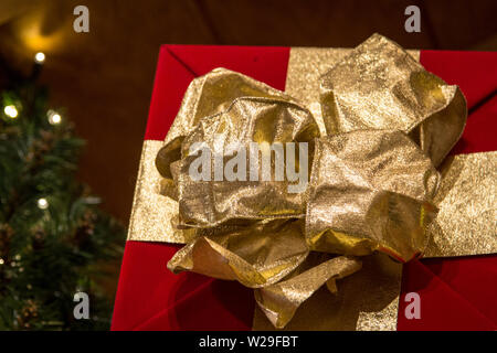 Shopping per il regalo di Natale perfetto. Avvolgere un regalo di Natale con la carta rossa e un ornato di prua in oro bianco con le luci di Natale in background Foto Stock