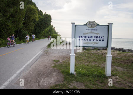 Mackinaw Island, Michigan, USA - Ciclisti su un'autostrada a due corsie intorno al perimetro dell'Isola di Mackinac con cartello del parco statale in primo piano. Foto Stock