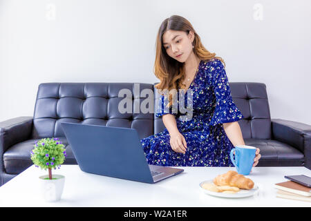 Giovane bella donna in vestito blu, prelievo di tazza di caffè mentre si lavora sul suo computer di casa sua soggiorno Foto Stock