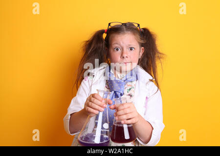 MR - esperimento scientifico a casa Geek Young Girl Età 7 - guardando pazzo a fotocamera tenere becher di scienza borosilicato pieno di sostanze chimiche sul giallo Foto Stock