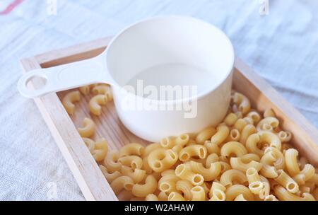 Il cibo e la cucina non cotti ed essiccato di pasta tipo gramigna o Pasta Gomiti nel vassoio in legno con bicchiere graduato in plastica. Foto Stock