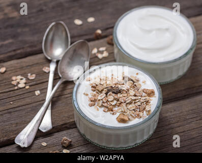 Yogurt bianco in recipiente di vetro con i fiocchi d'avena sulla parte superiore su rustiche scrivania in legno. Foto Stock