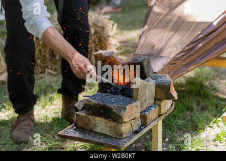 Mano con un pezzo di ferro riscaldato in un fuoco di carbone e dei soffietti fare faville dal fabbro strumenti. Il processo per la fabbricazione di utensili metallici e arma Foto Stock