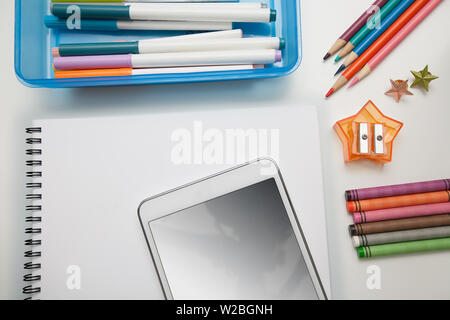 Scuola elementare di forniture con un dispositivo mobile, notebook, matite colorate, pastelli e pennarelli su una scrivania bianca. Foto Stock