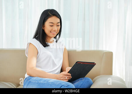 Sorridente piuttosto giovane donna asiatica in appoggio sul divano di casa e mediante applicazione sul computer tablet Foto Stock