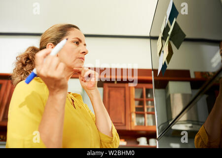 Malinconici donna matura rendendo lista della spesa e incollaggio note sul frigo prima di andare al supermercato Foto Stock