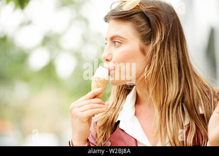 Ritratto di giovane donna bella mangiare gustoso gelato alla vaniglia nel cono di zucchero quando passeggiate all'aperto Foto Stock