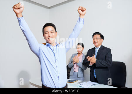 Emozionato felice giovane imprenditore asiatica i bracci di sollevamento e di celebrare la promozione dopo un incontro con la compagnia CEO e responsabile delle risorse umane Foto Stock