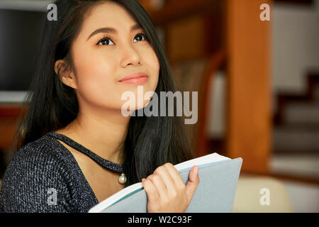 Giovane donna asiatica dreaming durante la lettura ha seduto con libro con malinconici occhi e guardando lontano Foto Stock