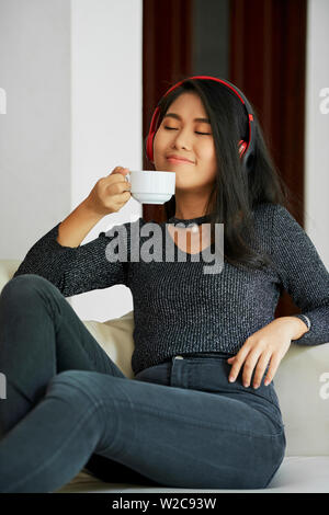 Giovane donna asiatica seduto sul divano godendo di tazza di caffè e ascolto di musica in cuffia senza fili Foto Stock
