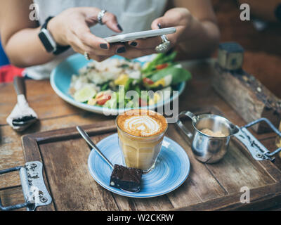 Donna di mani prendendo foto della tazza di caffè, Piccolo Latte, brownie con la colazione a tavola di legno da smartphone. Foto Stock