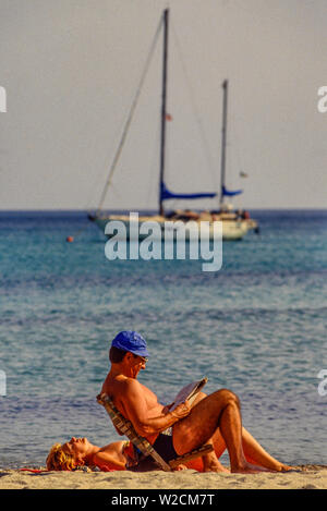 Spiaggia del Pevero, Sardegna, Italia. Un paio di prendere il sole. L'uomo legge un giornale. Barca ancorata nel mare dietro di loro. Foto: © Simon Grosset. Archivio: immagine digitalizzati da un originale di trasparenza. Foto Stock