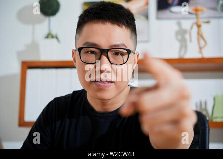 Ritratto di giovane uomo asiatico indossando occhiali puntando alla fotocamera e scegliendo lei mentre è seduto in ufficio Foto Stock