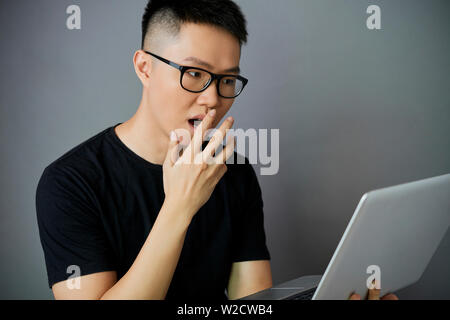 Asian giovane uomo in occhiali guardando il monitor del computer portatile e sorpreso isolate su uno sfondo grigio Foto Stock