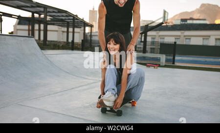 Felice giovane spingendo la sua ragazza seduta su skateboard a skate park. Giovane godendo allo skate park. Foto Stock