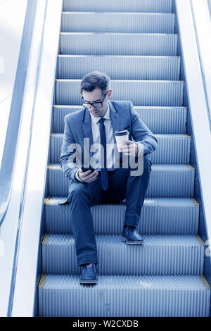 Imprenditore seduto su escalator navigando in rete e azienda caffè in mano Foto Stock