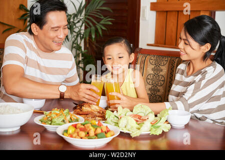 Felice famiglia asiatica seduti a tavola insieme con la loro figlia a cena e tostatura con bicchieri di succo d'arancia a casa Foto Stock