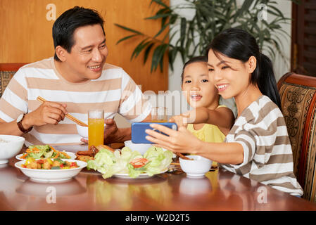 Sorridente giovane madre facendo una selfie con la sua famiglia sul telefono cellulare al tavolo durante la loro cena di famiglia a casa Foto Stock