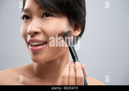 Close-up di Asian donna sorridente applicando toni crema sul suo viso con il make-up spazzola Foto Stock