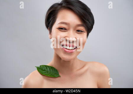 Ritratto di felice Asian giovane donna con corti capelli neri e con foglia verde sulla sua spalla e permanente di ridere Foto Stock