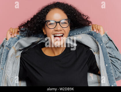 Colpo isolato di parentesi Allegra donna cerca su nuova camicia denim, indossa gli occhiali ottica, sorride ampiamente, ha perfetta atmosfera, pone contro la parete di rosa. Il pos Foto Stock