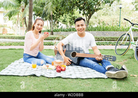 Ritratto di giovani asiatici Coppia sorridente alla telecamera mentre seduto sull'erba mangiare frutta e suonare la chitarra durante il picnic estivo nel parco Foto Stock