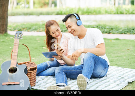 Felice coppia asiatica utilizzando tablet pc e il telefono cellulare sono seduti sul prato a guardare qualcosa e ridere durante il picnic nel parco Foto Stock