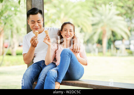Felice donna asiatica seduta sul banco e sorridente alla telecamera mentre a mangiare il gelato insieme con il suo fidanzato nel parco Foto Stock