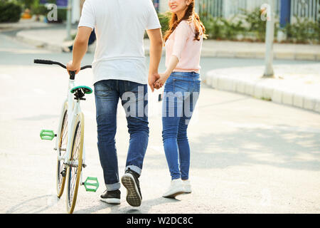 Vista posteriore del giovane coppia asiatica con passeggiate in bicicletta lungo la strada insieme e tenendo le mani Foto Stock