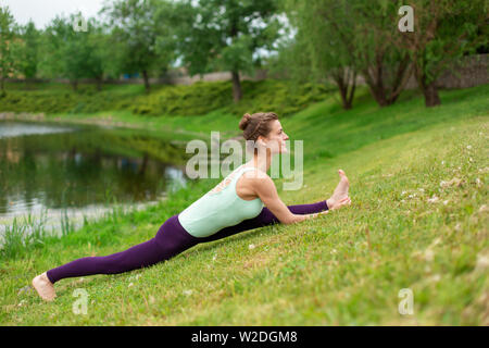 Slim bruna giovane ragazza yogi non difficile esercizi yoga sull'erba verde sullo sfondo dell'acqua. Foto Stock