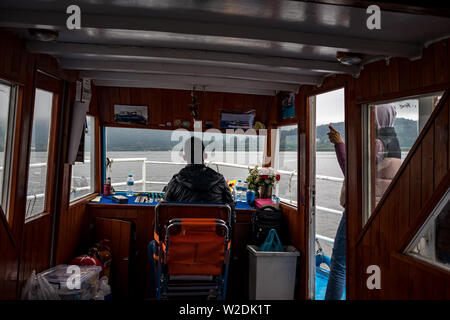 IOANNINA, Grecia - 6 giugno 2019 - Maschio capitano regole vecchie di piccole dimensioni di colore bianco e rosso crociera turistica traghetto sul lago Pamvotida vicino alla piccola e bella città greca. Vista posteriore interno della cabina. Mattinata nebbiosa Foto Stock