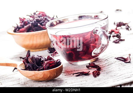 Primo piano alla ciotola di legno tazza da tè e un cucchiaio di ibisco secco petali su tela di lino sfondo Foto Stock