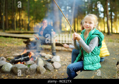Carino piccole sorelle e il padre loro tostatura di marshmallows su bastoni al falò. Bambini che si divertono a camp fire. Il campeggio con i bambini rientrano nella foresta. F Foto Stock