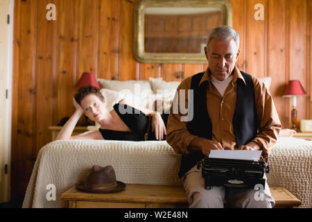 Uomo maturo utilizzando una macchina da scrivere nella camera da letto mentre sua moglie giace sul letto dietro di lui. Foto Stock